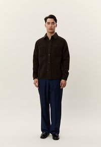 Jaunas vyras stovi, dėvi tamsiai rudą užsegamą striukę su krūtinės kišenėmis, tamsiai mėlynas kelnes ir juodus batus, priešais vienspalvį foną.