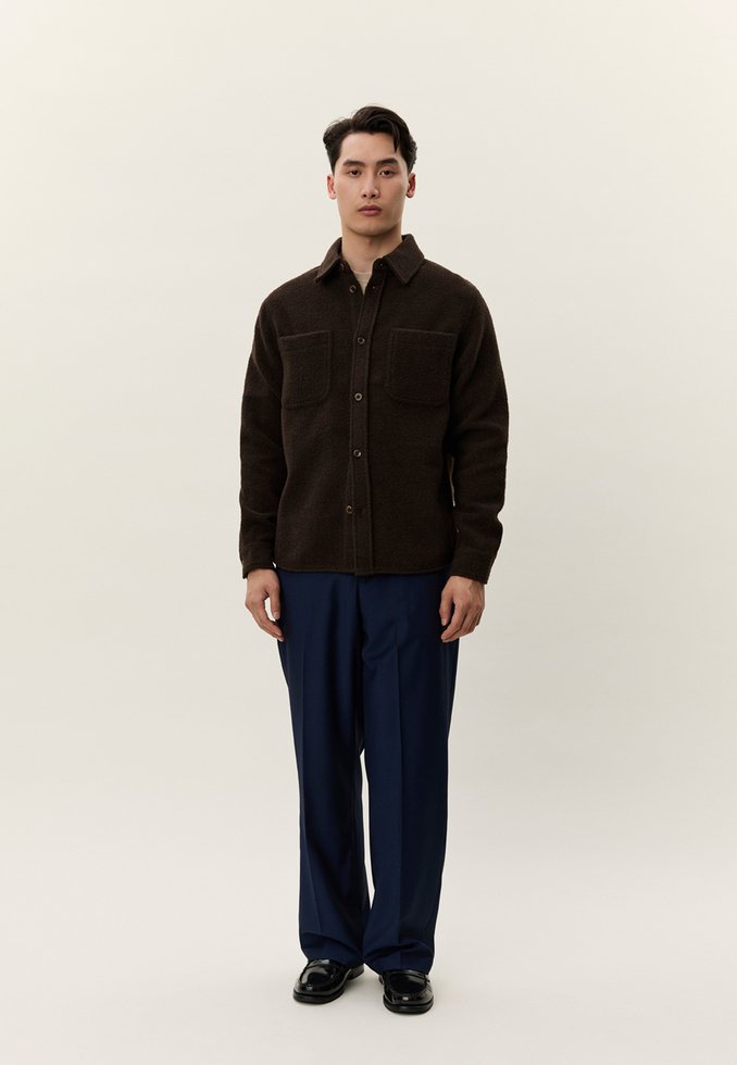 Jaunas vyras stovi, dėvi tamsiai rudą užsegamą striukę su krūtinės kišenėmis, tamsiai mėlynas kelnes ir juodus batus, priešais vienspalvį foną.