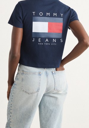 Persoon draagt een marineblauw cropped t-shirt met een rood, wit en blauw logo en lichtblauwe hooggetailleerde jeans, hand in achterzak.