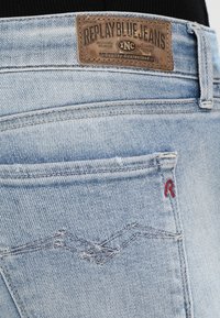 Jasnoniebieskie dżinsy z wyhaftowanym tylnym kieszonką, małym czerwonym haftem "R" oraz skórzaną metką marki "Replay Blue Jeans."