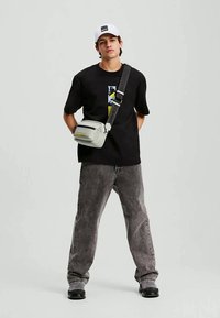 Jovem vestido com uma t-shirt gráfica preta, jeans cinzentos, sapatos pretos, boné branco e uma bolsa crossbody cinza claro com uma alça larga.