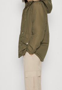 Olivově zelená péřová bunda s odnímatelnou kapucí s kožešinou, na zip a s bočními kapsami, v kombinaci se světlými béžovými cargo kalhotami.