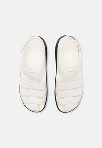 Paire de sandales blanches à bout fermé avec semelles noires, comprenant plusieurs brides et une sangle arrière pour un maintien sûr, vues de dessus.