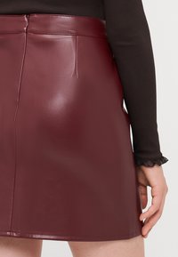 Burgundské mini sukne z faux kože s hladkým povrchom, priliehavým strihom, detailmi na zadnom šve a vysokým pásom. Akcentované šitím.