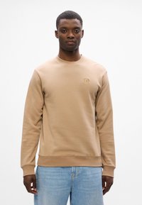 Felpa marrone con collo rotondo, maniche lunghe e polsini a costine. Presenta un piccolo logo in rilievo sul petto. Abbinata a jeans azzurri chiari.