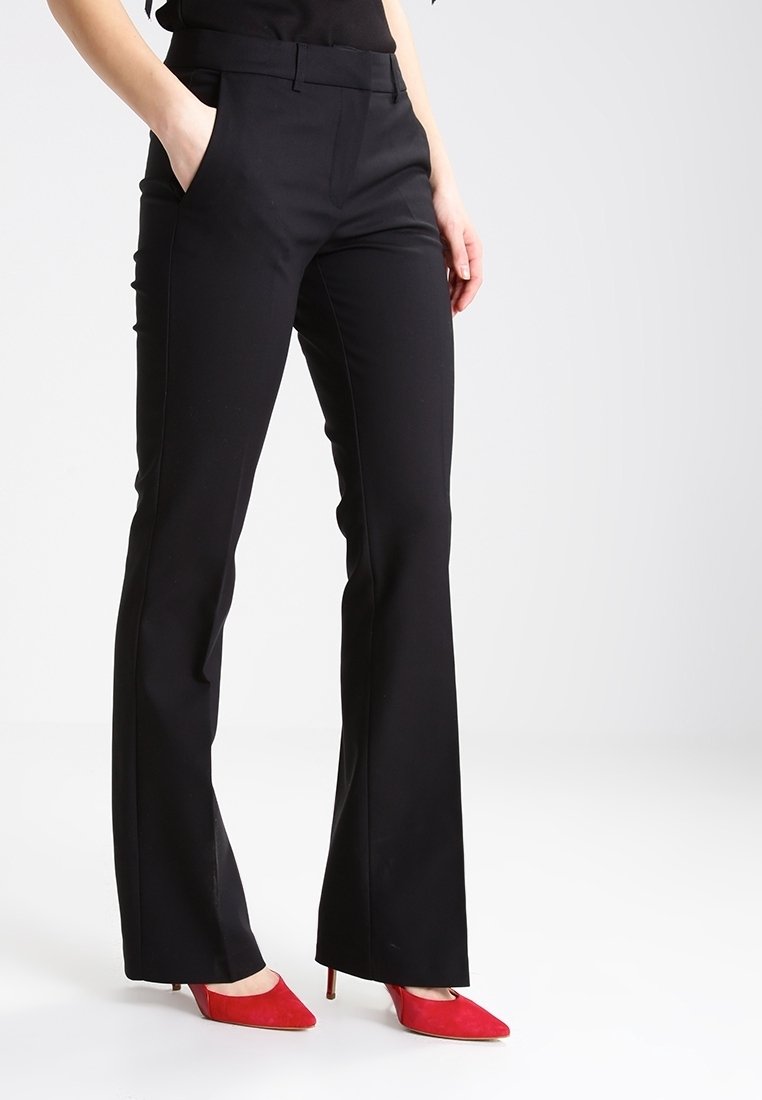 Personne portant un pantalon noir sur mesure et des chaussures à talons rouges, debout avec une main dans la poche sur un fond uni.