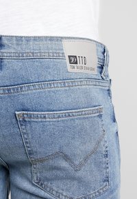 Světlé modré džínové kalhoty s zadním kapsou se všitým vzorem šipky a šedou etiketu s nápisem „TTD TOM TAILOR DENIM BRAND“.