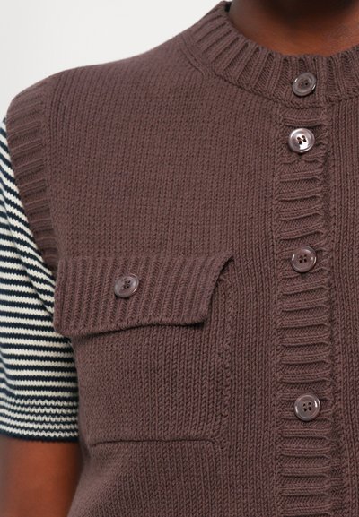 Gilet lavorato a maglia marrone con colletto alto, dotato di chiusura a bottoni, tessuto testurizzato e taschino sul petto con bottone. Camicia a righe sotto.