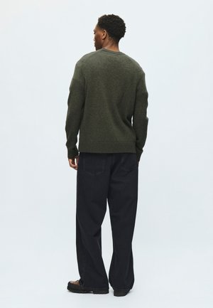 Homme de dos portant un pull vert foncé, un pantalon large noir et des chaussures marron sur un fond clair uni.