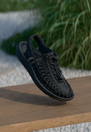 Sandalo flessibile nero con design superiore intrecciato, dotato di un sistema di lacci sicuro e di una suola imbottita. Posato su una superficie di legno.