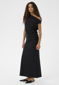 Kvinde med langt krøllet hår iført en sort asymmetrisk maxi-kjole med rynker og sorte spidse flade mules, stående foran en hvid baggrund.