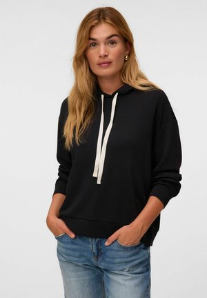 Young woman with long blonde hair wearing a black hoodie with white drawstrings and blue jeans, standing with hands in pockets.