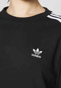 Svart bomullströja med rund halsringning, med en vit Adidas-logotyp och tre vita ränder på varje axel. Mjuk textur.