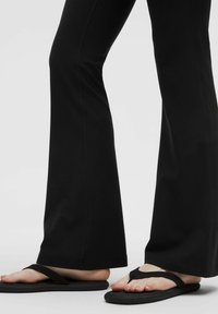 Schwarze Schlaghosen mit glatter Textur, kombiniert mit schwarzen Flip-Flops mit flachen Sohlen und minimalistischen Riemen.