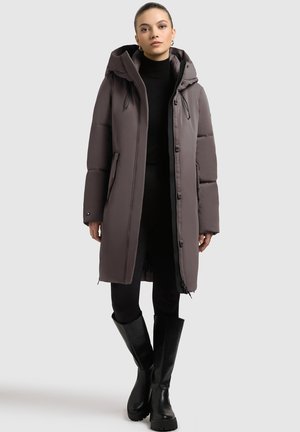 Duga, kapuljača, izolirana parka u tamnoj taupe boji s elegantnim dizajnom, bočnim džepovima i kontrastnim crnim detaljima. Nosi se preko crnog topa i hlača.