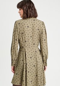 Robe vert olive avec de longues manches bouffantes et une taille cintrée. Présente un motif floral et de feuilles en noir sur l'ensemble du tissu.