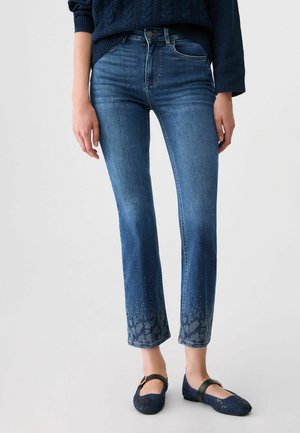 Femme portant un jean slim bleu avec un motif floral aux poignets, un pull bleu foncé et des chaussures bleu foncé assorties avec des brides à la cheville.