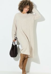 Abito maglione beige lavorato a maglia con dettagli a costine, maniche lunghe e vestibilità rilassata, abbinato a una borsa nera intrecciata e stivali al ginocchio color caramello.