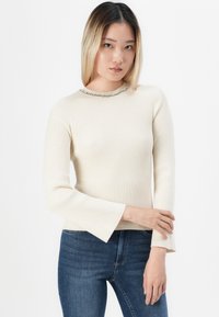 Young woman with straight blonde hair wears a cream ribbed sweater with beaded neckline and blue jeans, posing against a white background.