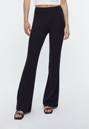 Femme portant un pantalon noir flare et des sandales noires à brides ouvertes à l’orteil, avec un débardeur blanc court visible au-dessus de la taille.