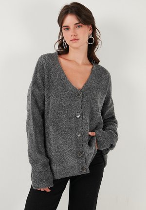 Cardigan grigio in peluche con scollatura profonda a V, grandi bottoni e maniche oversize, realizzato in un tessuto morbido e testurizzato. Pantaloni neri sotto.
