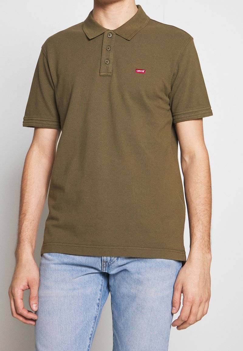 Homme portant un polo à manches courtes vert olive avec trois boutons et un petit logo rouge Levi's, associé à un jean bleu clair.