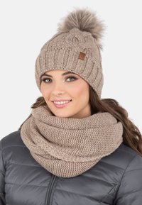 Bonnet en tricot beige avec un pompon en fourrure, présentant une texture côtelée et un patch logo. Accompagné d'une écharpe infinity assortie et confortable.