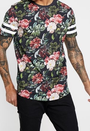 Chemise noire à manches courtes avec des rayures blanches sur les manches, couverte de grands motifs floraux rouges, roses et blancs ; la personne a les bras tatoués.