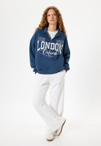 Sudadera con cremallera en azul marino con el gráfico "LONDON Oxford" en blanco, combinada con pantalones acampanados blancos y zapatillas crema. Textura de tela suave.