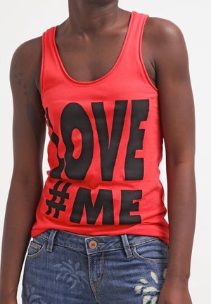 Personne portant un débardeur rouge avec un grand texte noir "LOVE #ME" et un jean bleu avec un motif floral clair.