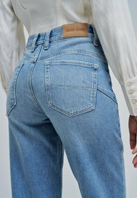 Jeans Salsa de un azul claro con bolsillos traseros y una etiqueta de marca de cuero marrón, combinados con una camisa blanca de manga larga.