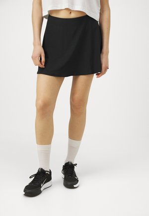 Person, die einen schwarzen kurzen Rock, weiße Socken, schwarze Turnschuhe und ein kurzes weißes Top trägt, vor einem einfarbigen weißen Hintergrund steht.