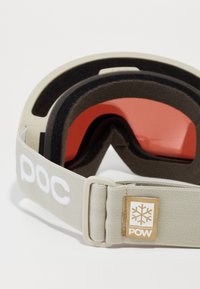 Beige szíjjal, piros lencsével, puha habpárnázattal rendelkező hópárás szemüveg, hópihe logóval ellátott márkás címkével.