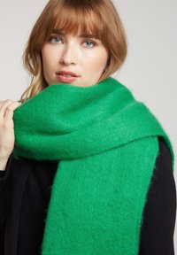 Écharpe verte en matériau doux et texturé, drapée autour du cou, avec un design épais et des bords légèrement effilochés.