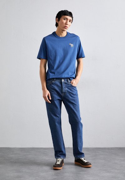 Mėlynas medvilninis marškinėlis su maža grafika, derinamas su klasikiniais mėlynais džinsais ir tamsiais sportbačiais su baltais raišteliais bei ruda padu.