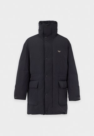 Homme portant un bonnet gris, une veste noire, un pull bleu marine avec logo, un pantalon noir et des chaussures noires, debout, une main dans la poche, regardant de côté.