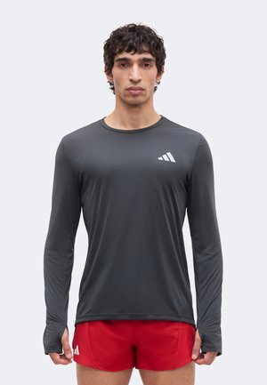 Man draagt een donkergrijs langarmig sportshirt en rode korte broek, staat recht vooruit gericht met een neutrale uitdrukking tegen een effen achtergrond.