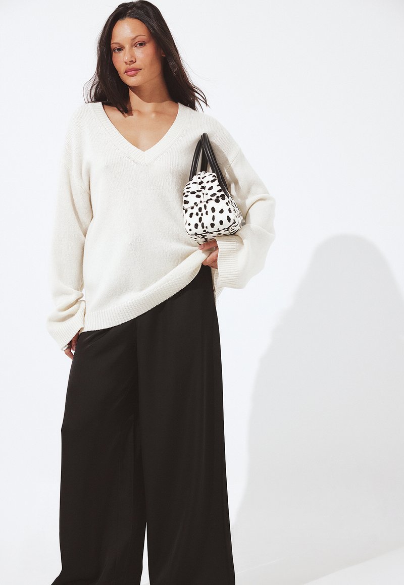 Donna che indossa un ampio maglione bianco a V e pantaloni neri larghi, che tiene sulla spalla una borsa bianca con macchie nere.