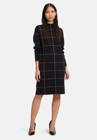 Svart stickad klänning med orange rutmönster, hög krage, långa ärmar, rak skärning och faller precis ovanför knäna. Bärs med svarta klackar.