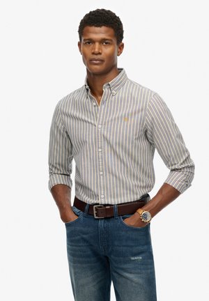 Gestreiftes Button-Down-Hemd aus hellblauem und beigem Baumwollstoff, kombiniert mit dunklen Jeans. Ein brauner Ledergürtel und eine Uhr runden das Outfit ab.