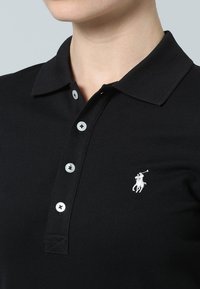 Schwarzes Poloshirt mit klassischem Kragen, Dreiknopfleiste und kleinem weißem Logo auf der linken Brustseite. Hergestellt aus strukturiertem Stoff.