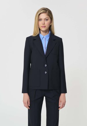 Femme aux cheveux blonds raides portant un blazer bleu marine par-dessus une chemise bleu clair boutonnée, sur un fond uni clair.