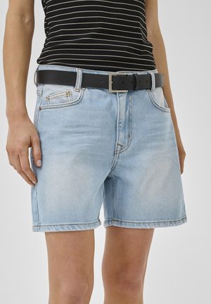 Egy személy világoskék farmer rövidnadrágot visel, fekete övvel és fekete-fehér csíkos ujjatlan felsővel.
