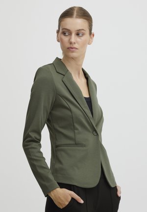 Woman with pulled-back hair wearing an olive green fitted blazer over a black top, hands in pockets, looking to her left against plain background.