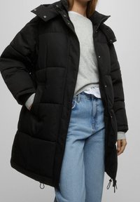 Doudoune noire avec capuche amovible, fermeture éclair à l'avant et boutons-pression. Présente des détails matelassés et des poches latérales. Portée par-dessus un pull gris et un jean.