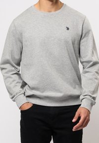 Man som bär en ljusgrå långärmad tröja med en liten mörk logotyp på bröstet, med händerna i fickorna på svarta byxor, visas från nacken till höfterna.