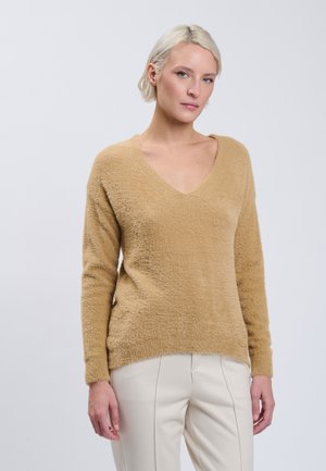 Lyshåret kvinde iført en beige fuzzy V-hals sweater og cremefarvede bukser, stående foran en ensartet lysegrå baggrund.