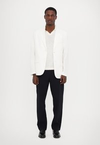 Weißes Leinenblazer über einem grauen Pullover, kombiniert mit marineblauen Hosen und schwarzen Schuhen. Das Blazer hat zwei Knöpfe und minimale Designdetails.