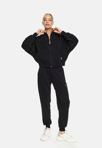 Schwarze cropped Zip-Jacke mit überschnittenen Schultern und Taschen, kombiniert mit passenden taillierten Sweatpants. Beide Teile tragen dezente Markenlogos. Weiße Sneakers.