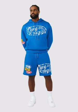 Mann steht mit den Händen hinter dem Rücken und trägt einen leuchtend blauen Hoodie sowie passende Shorts mit weißen und mehrfarbigen True Religion-Logos, weiße Sneakers.